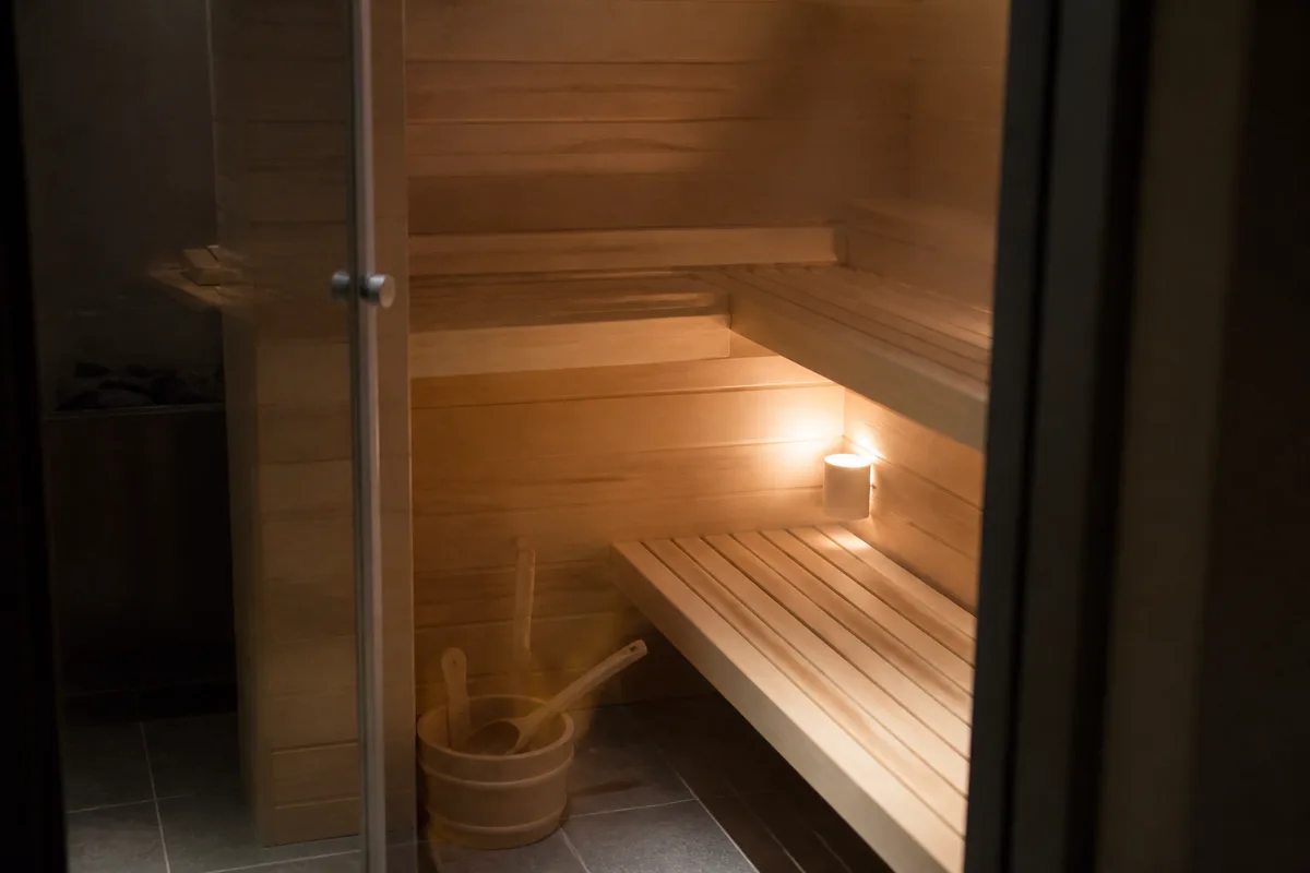 Petit sauna en bois clair vu par la porte vitrée, lumière chaude sur les banquettes